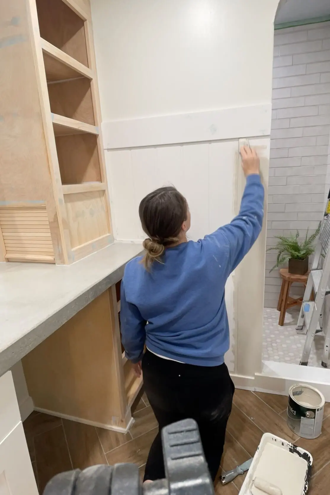 A woman in a blue sweatshirt applying spackle to a newly installed white shiplap wall in a bathroom renovation. The scene features a concrete countertop with an unfinished wooden cabinet and an arched shower entrance with brass fixtures in the background.